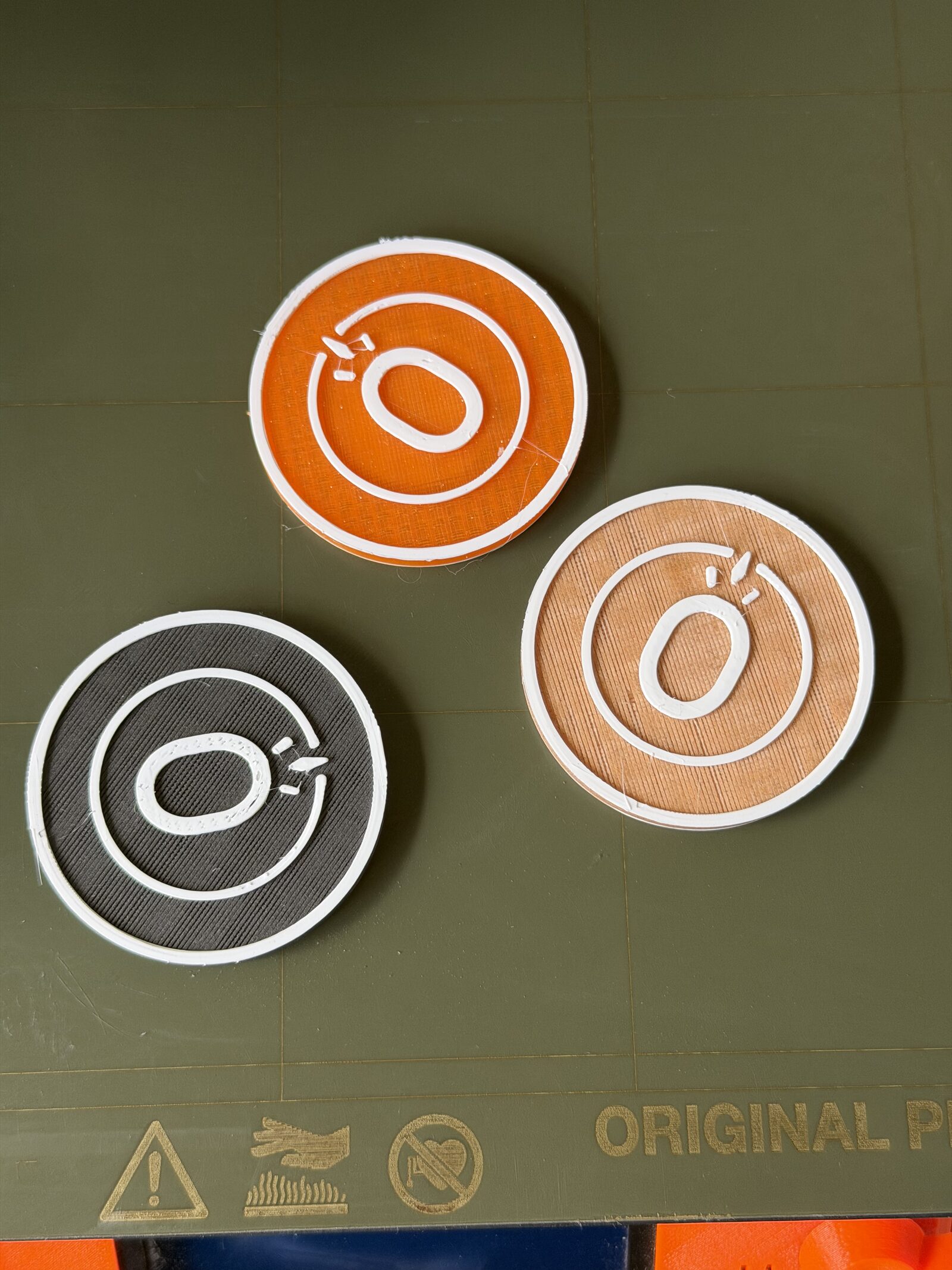 Three 3D-printed Block Coins in clay orange, copper, and ink black on a printer bed, each with a raised ring-and-zero emblem.